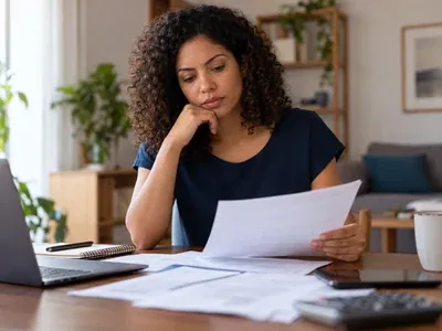 Mulher com a mão no queixo avaliando papéis de contrato ao lado do notebook